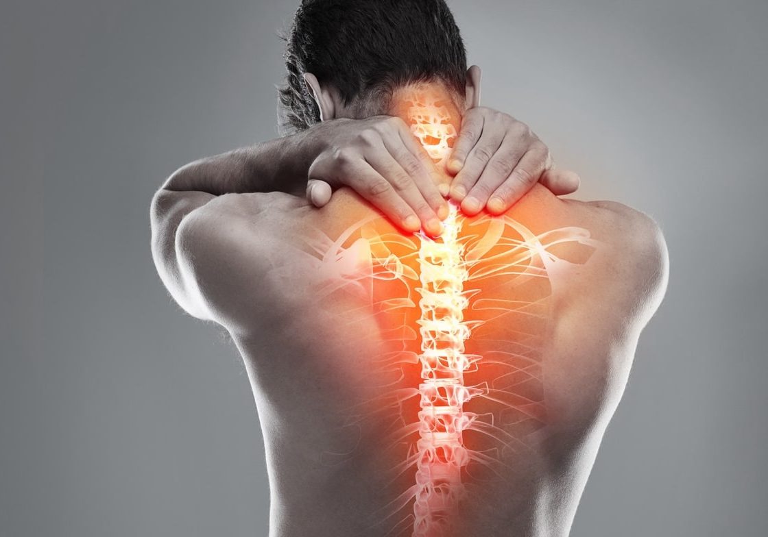 A person seen from behind holds their neck and upper back in discomfort. An illustration of the spine is superimposed, glowing red to indicate pain or inflammation along the spinal column.