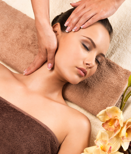 A woman with closed eyes relaxes on a brown towel while receiving a gentle head and neck massage. Yellow flowers are placed nearby, creating a calm, spa-like atmosphere.